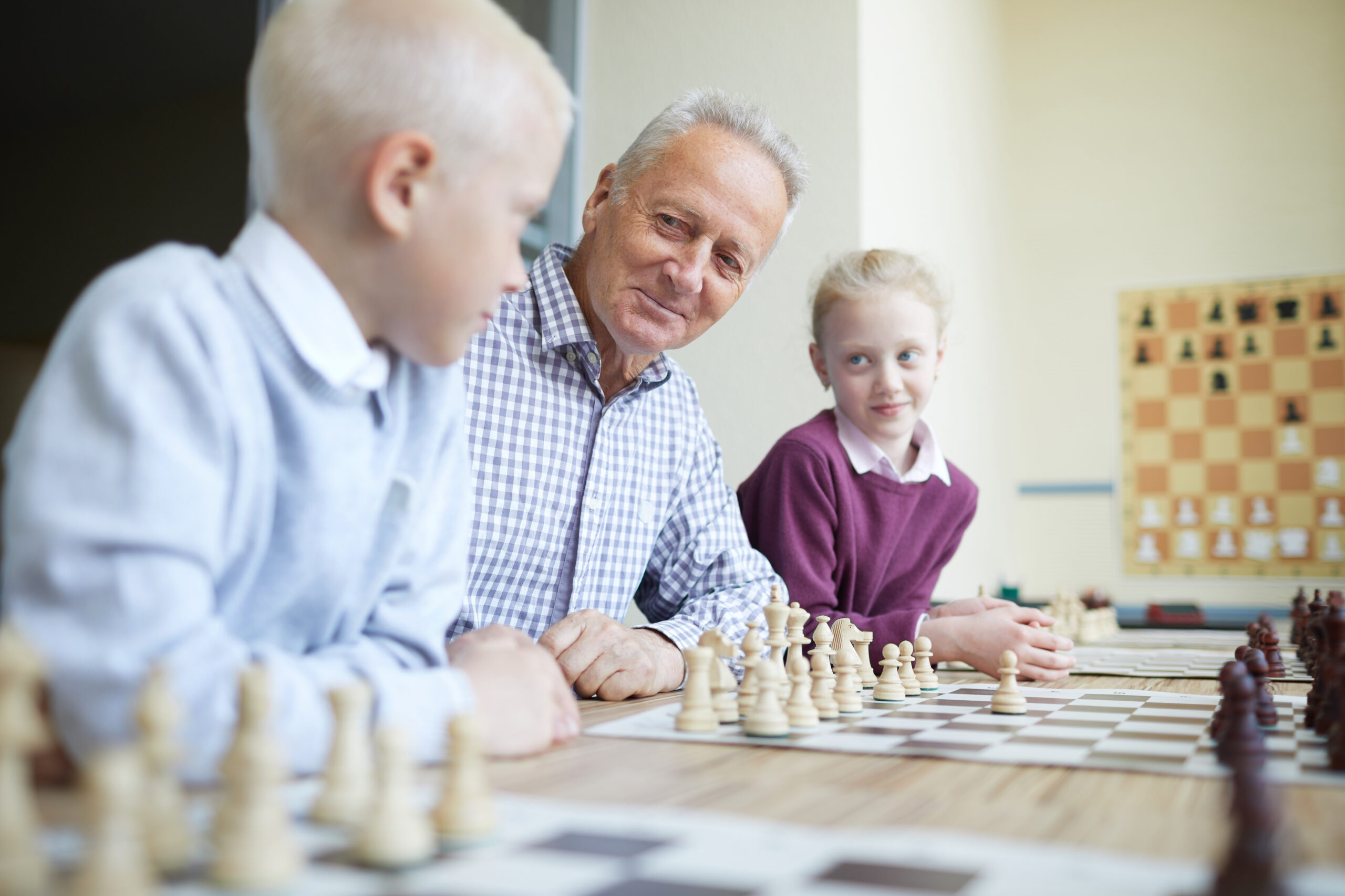 Smiling grandfather in checked shirt teaching his two curious grandkids how to play chess