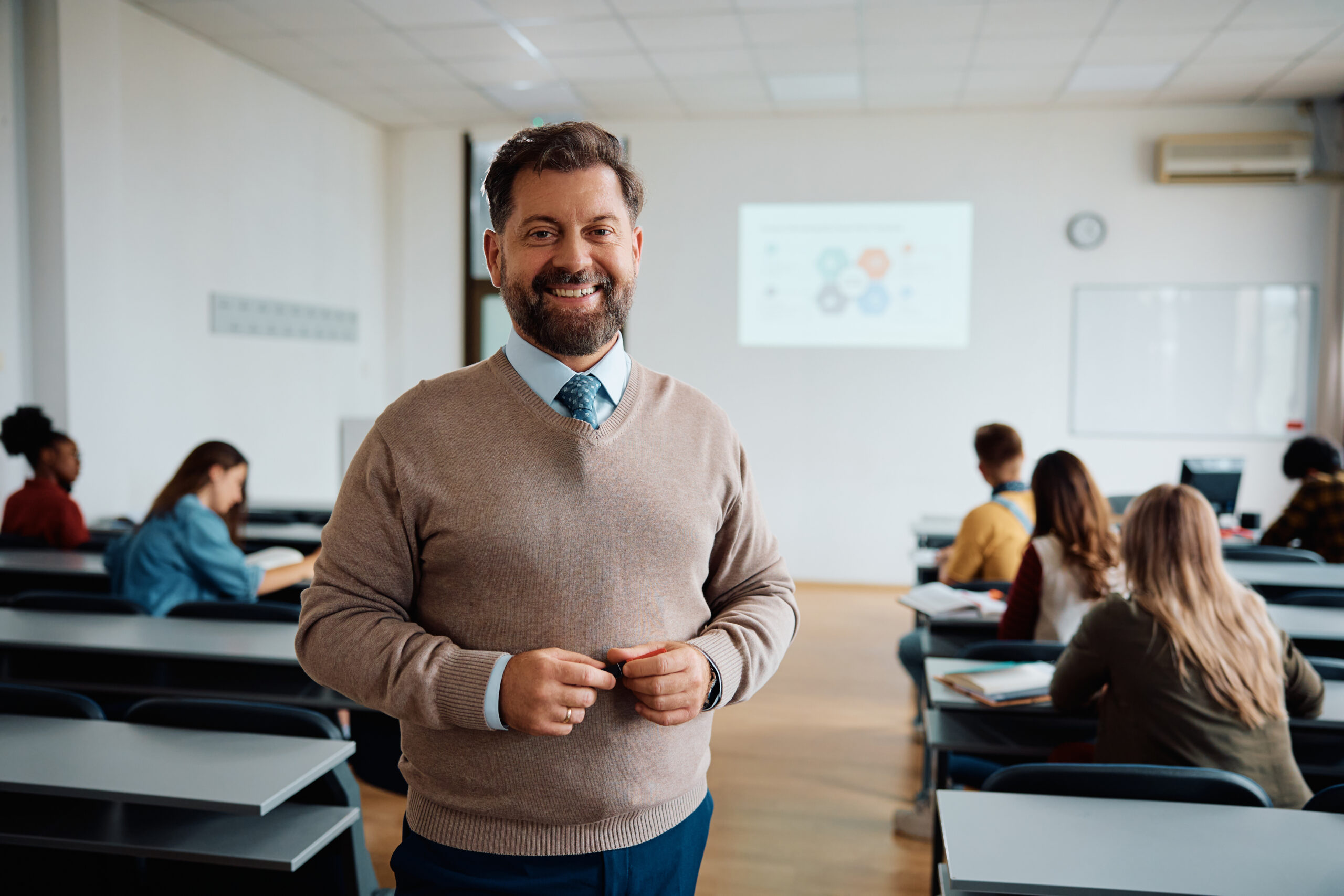 Happy professor in lecture hall at the university looking at camera. His students are int he background.